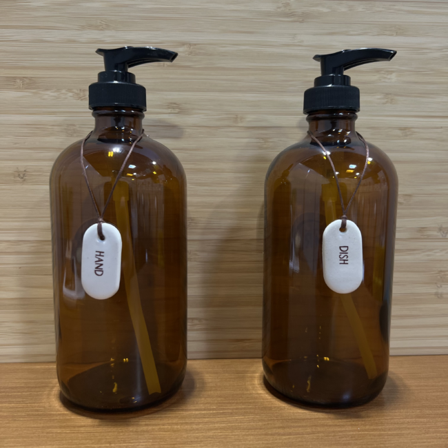 Two amber glass bottles with pump tops - each with a white ceramic tag around the bottle's neck. One reads "Hand" and one reads "Dish."