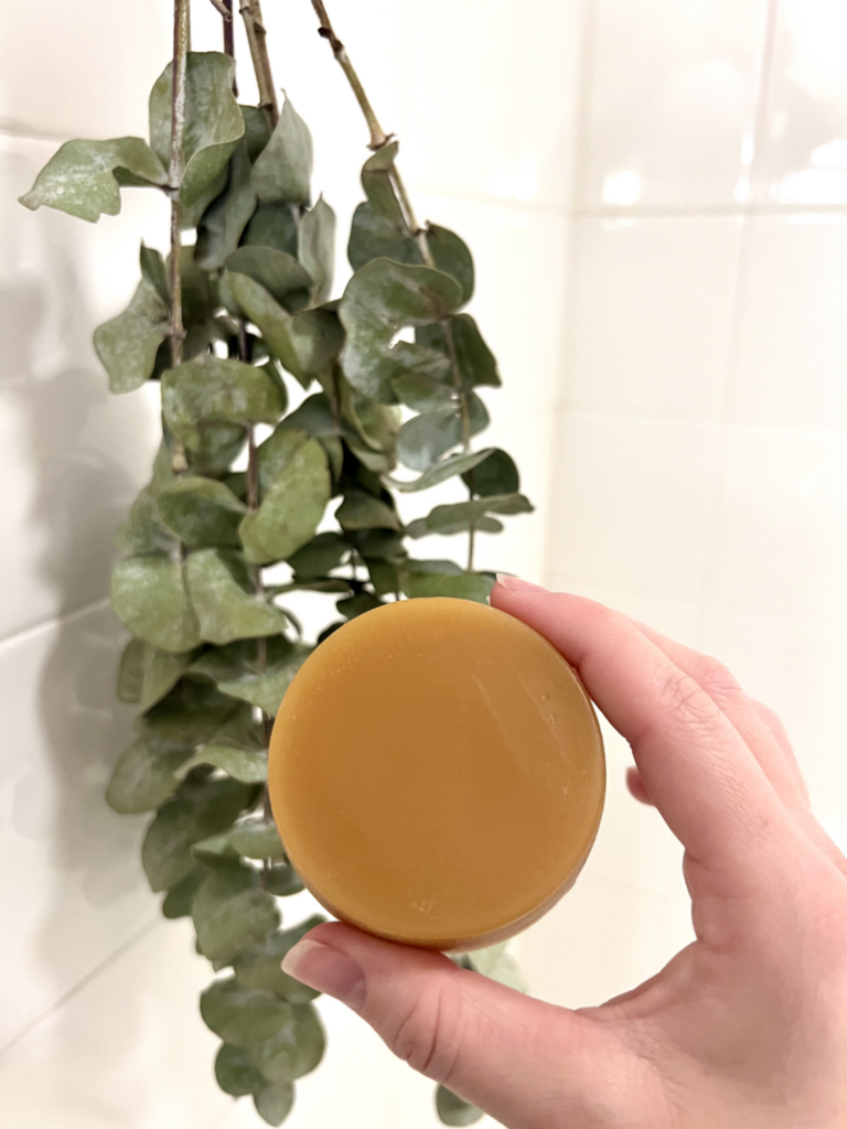 A hand holding a full-sized conditioner bar. In the faded background is a white tile shower with eucalyptus.